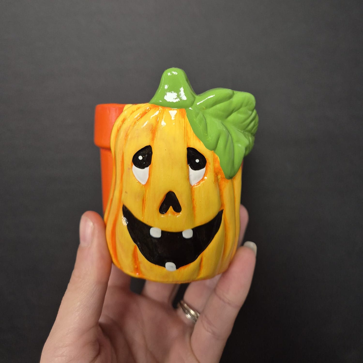 Hand holding a small pumpkin-shaped container with a face against a dark background